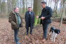 drei Männer stehen im Wald auf braunem Laub, ein schwarz-weißer Hund steht neben dem größten Mann