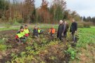 5 Menschen knien in einem Acker, drei Männer stehen vor ihnen, auf dem Acker sind kleine Eichenplanzen