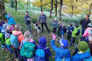 Kinder stehen im Kreis im Wald, Mann mit Hund begrüßt die Kinder, ein Mann hält eine Holzscheibe