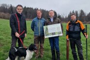 4 Männer und ein Hund stehen auf einer Wiese. Ein Mann hält eine große Tafel vor seiner Brust