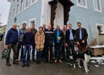 eine große Gruppe Personen steht vor dem Eck eines blauen Hauses, ein Mann hat einen schwarzweißen Hund an der Leine