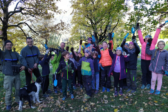 Eine Gruppe mit vielen Kindern steht im Wald, links zwei Männer im Vordergrund ein Hund