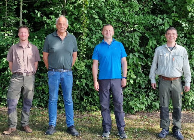 vier Männer stehen in einer Reihe mit dem Gesicht zur Kamera, im Hintergrund eine Laubhecke