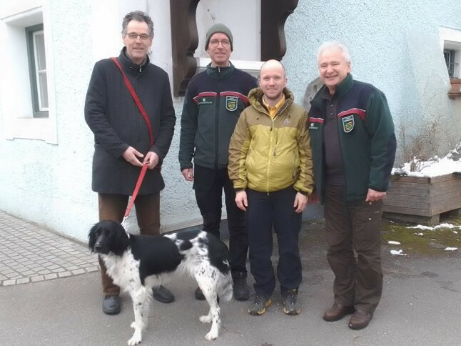 Auf dem Foto stehen vier Männer vor der Fassade eines hellblauen Gebäudes. Der Mann links im Bild führt einen schwarz-weiß gefleckten Hund an einer roten Leine mit sich