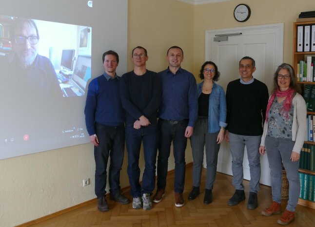 6 Personen stehen in einem Raum vor einer Leinwand, auf der Leinwand sitzt ein Mann mit Bart und Brille vor seinem Computer und schaut in die Kamera